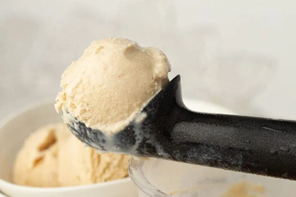 A close-up of a black ice cream scoop holding a scoop of beige ice cream, with a bowl of more ice cream in the background.