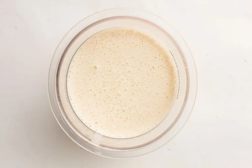 A clear glass filled with a frothy, light beige liquid seen from above on a plain white surface.