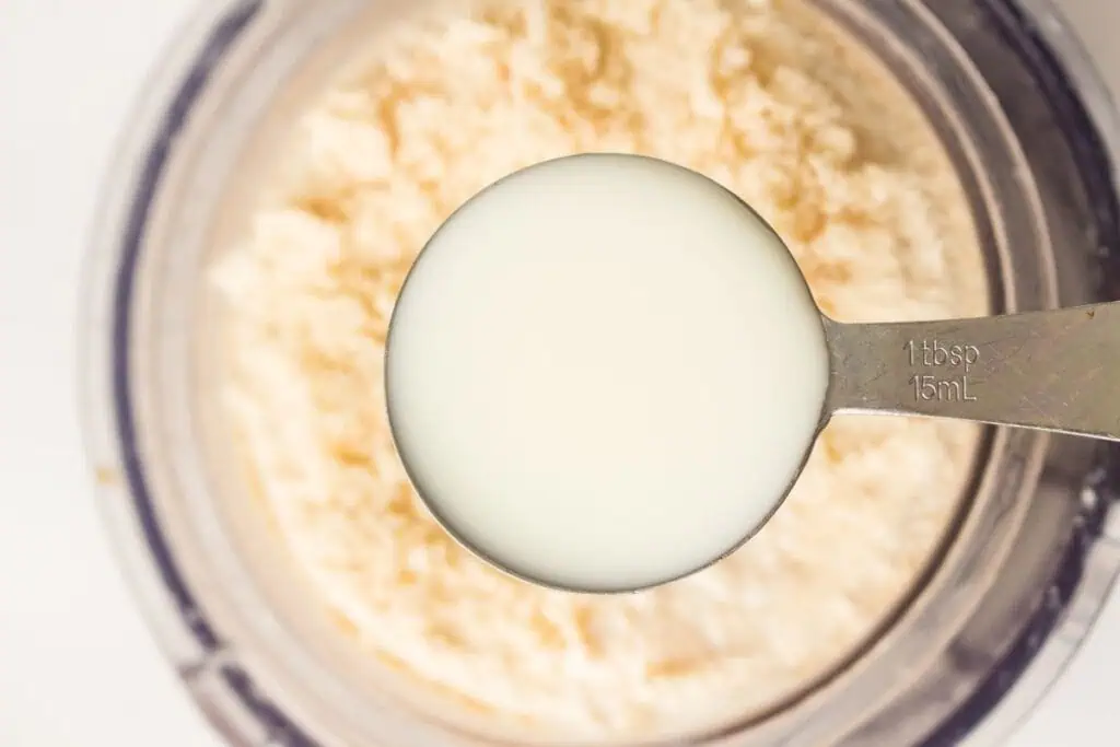 A metal tablespoon filled with milk is held above a bowl containing a dry, crumbly mixture.