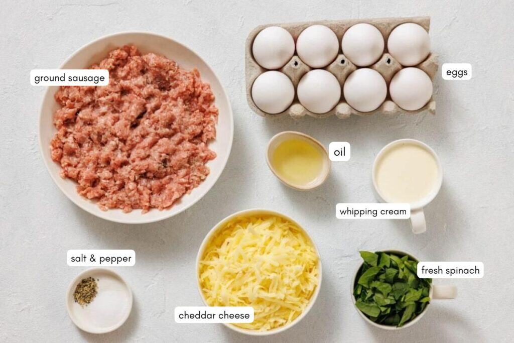 Bowls of ground sausage, shredded cheddar cheese, fresh spinach, salt and pepper, a carton of eggs, a cup of oil, and a cup of whipping cream are arranged on a light surface.