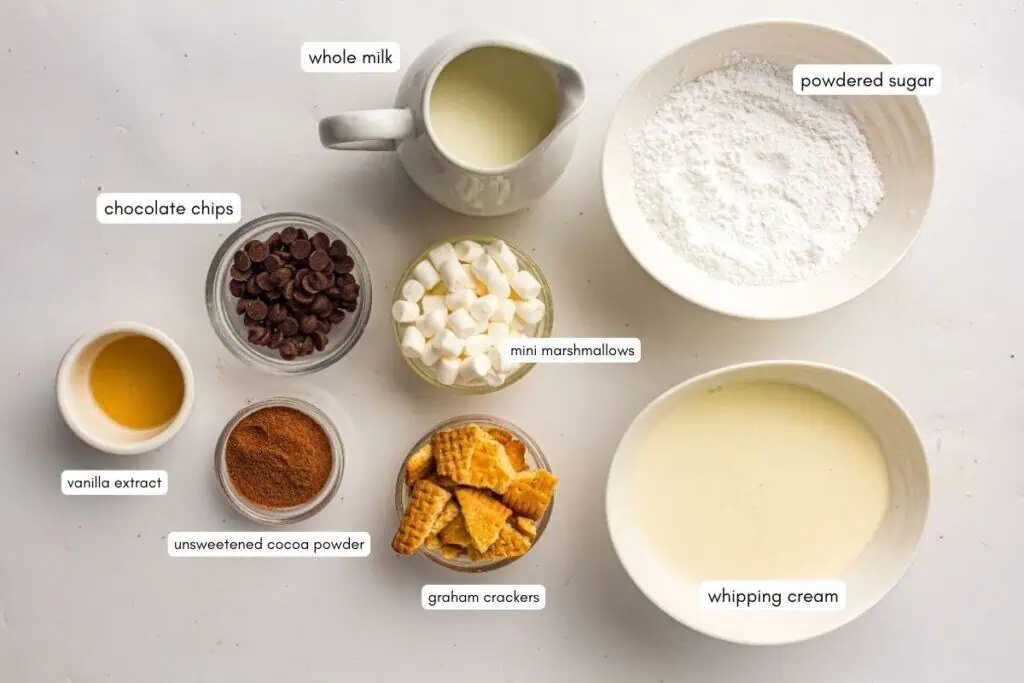 Top-down view of bowls and a pitcher containing whole milk, powdered sugar, chocolate chips, mini marshmallows, vanilla extract, unsweetened cocoa powder, graham crackers, and whipping cream.