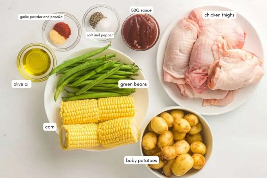 Various labeled ingredients on a white surface: raw chicken thighs, green beans, corn, baby potatoes, olive oil, BBQ sauce, garlic powder, paprika, salt, and pepper.