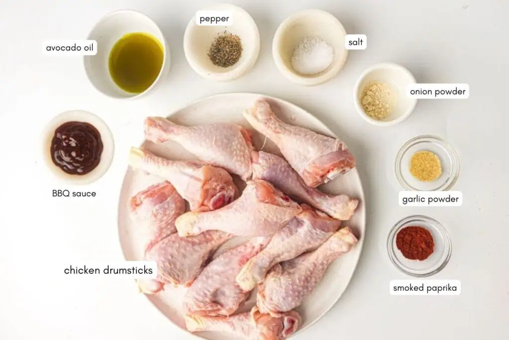 Raw chicken drumsticks on a plate surrounded by small bowls containing BBQ sauce, avocado oil, pepper, salt, onion powder, garlic powder, and smoked paprika. Each ingredient is labeled.