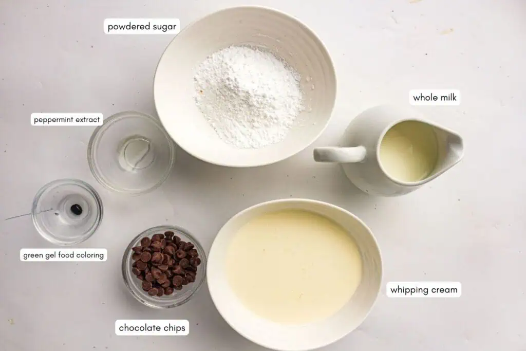 Top-down view of bowls and jars containing powdered sugar, whole milk, whipping cream, chocolate chips, green gel food coloring, and peppermint extract, each labeled.