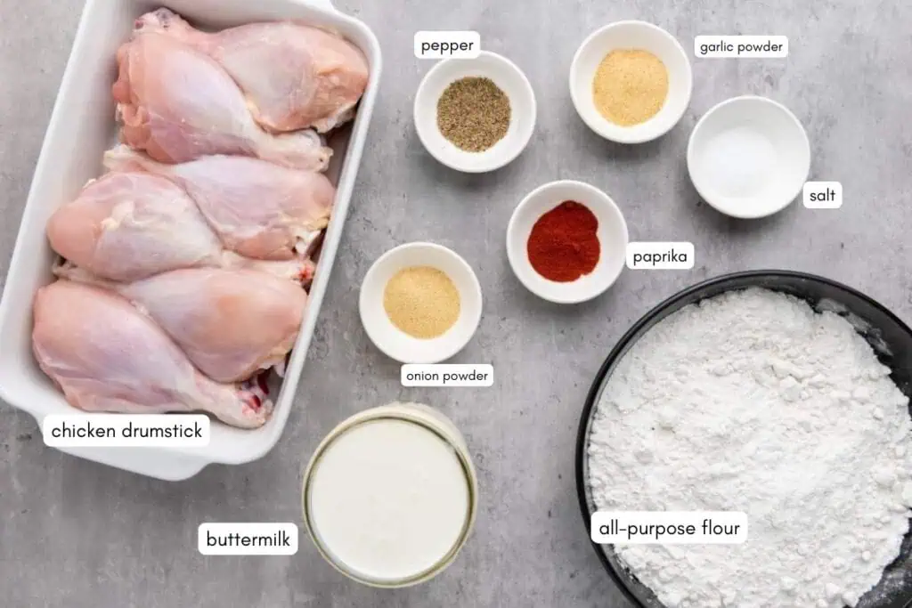 Raw chicken drumsticks, all-purpose flour, buttermilk, and small bowls containing onion powder, garlic powder, paprika, salt, and pepper are arranged on a gray surface.
