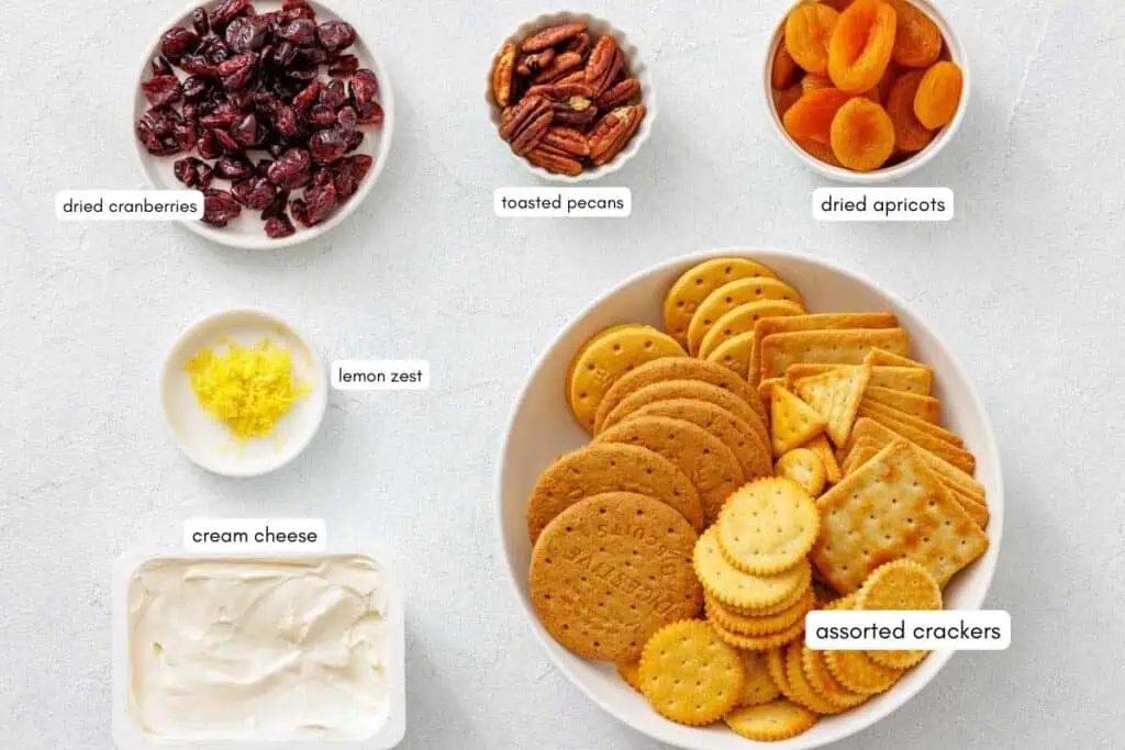 Bowls of dried cranberries, toasted pecans, dried apricots, lemon zest, cream cheese, and assorted crackers arranged on a white surface.