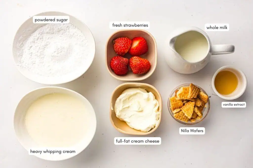 Bowls containing powdered sugar, fresh strawberries, whole milk, heavy whipping cream, full-fat cream cheese, vanilla extract, and broken Nilla Wafers are arranged on a white surface.