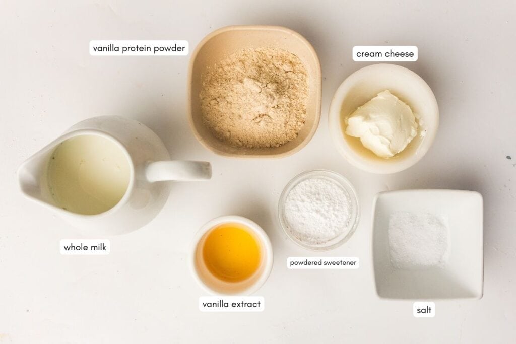 Six labeled ingredients on a white surface: vanilla protein powder, cream cheese, whole milk, vanilla extract, powdered sweetener, and salt.