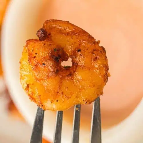 A seasoned, cooked shrimp on a fork is held above a bowl of dipping sauce.