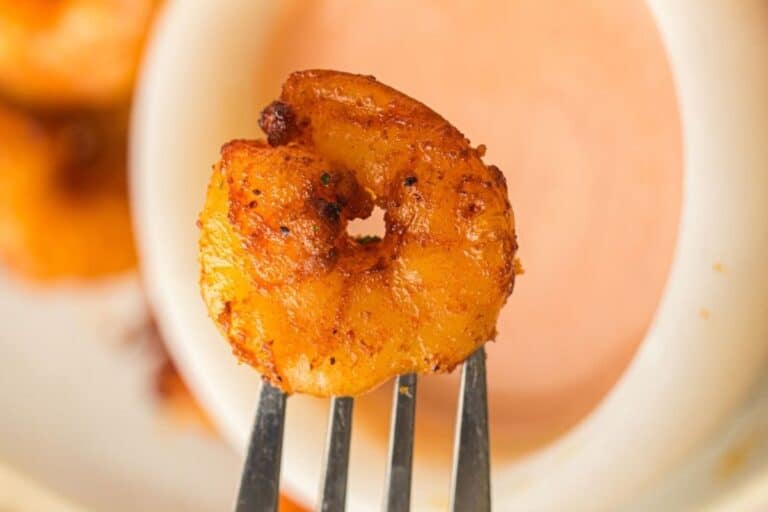 A seasoned, cooked shrimp on a fork is held above a bowl of dipping sauce.