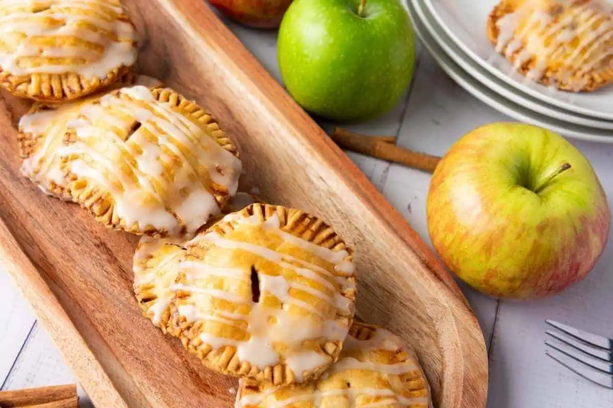 Four small glazed apple hand pies are arranged on a wooden tray, with whole apples and stacked white plates in the background.