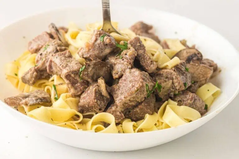 A white bowl filled with wide egg noodles topped with chunks of beef in a creamy sauce, with a fork sticking into the pasta.