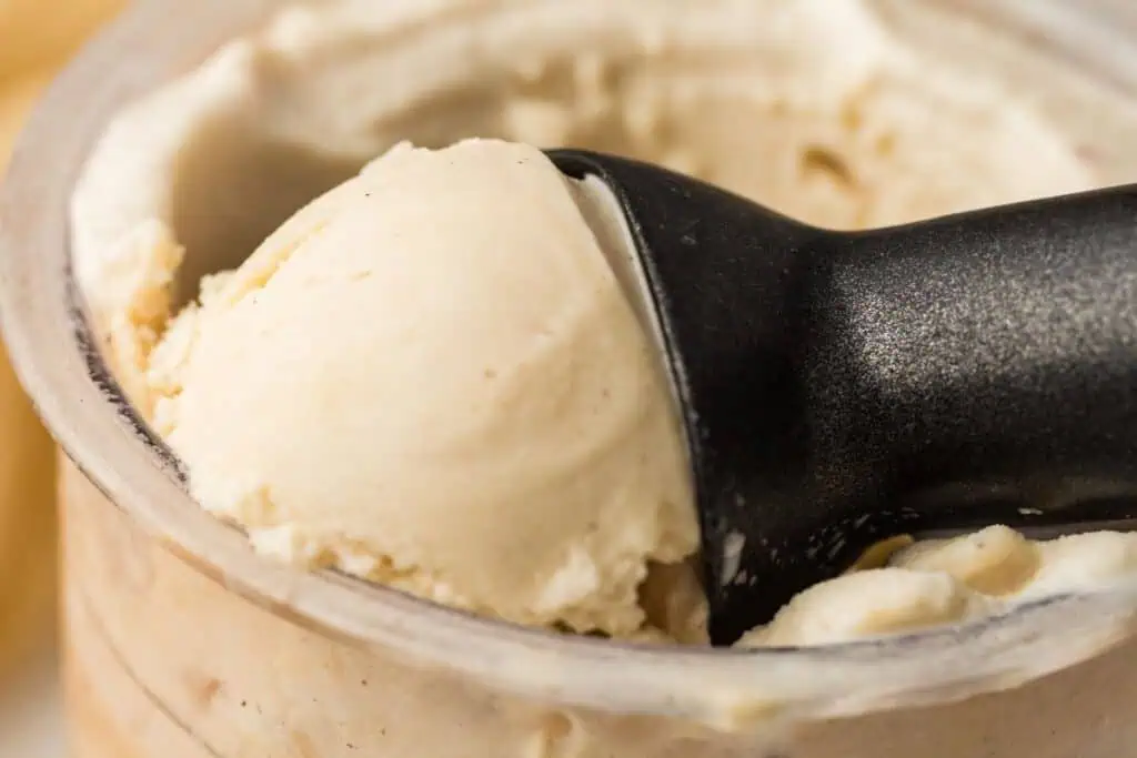 Close-up of a black ice cream scoop lifting a portion of vanilla ice cream from a container.