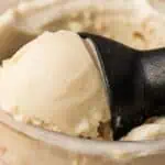 Close-up of a black ice cream scoop lifting a portion of vanilla ice cream from a container.