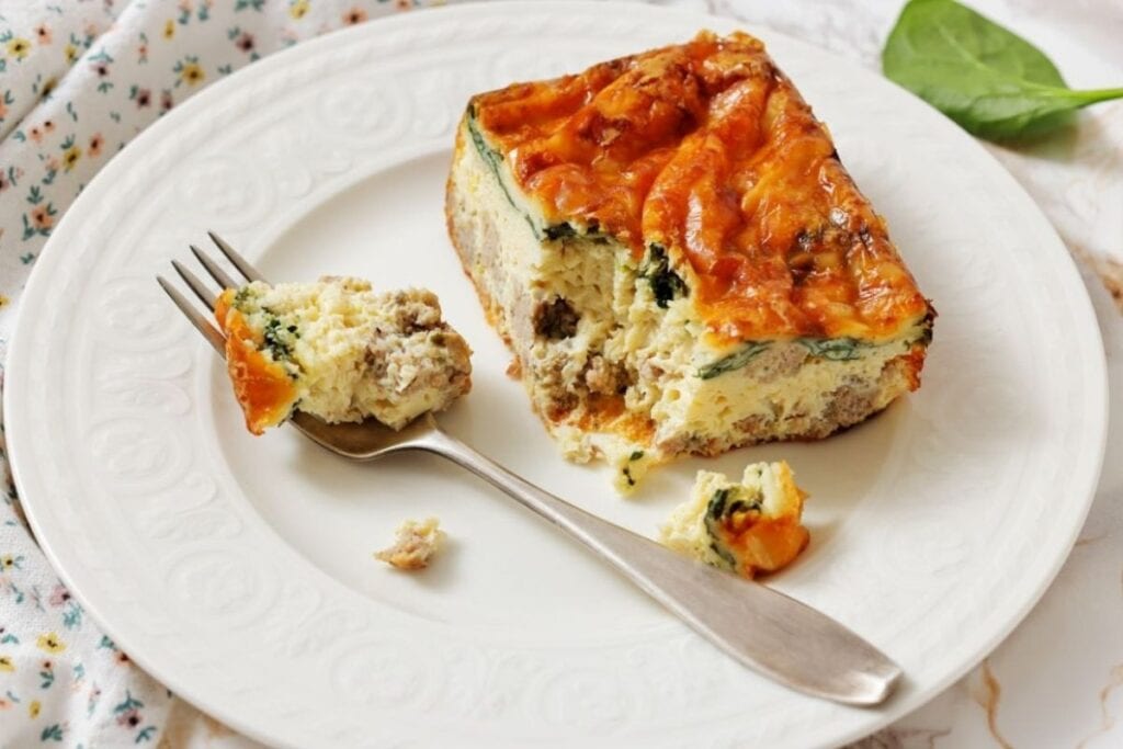 A slice of baked egg casserole with spinach and sausage on a white plate, with a fork holding a small bite.