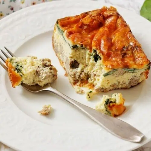 A slice of Sausage, Spinach, and egg casserole on a white plate, with a fork holding a bite-sized piece.