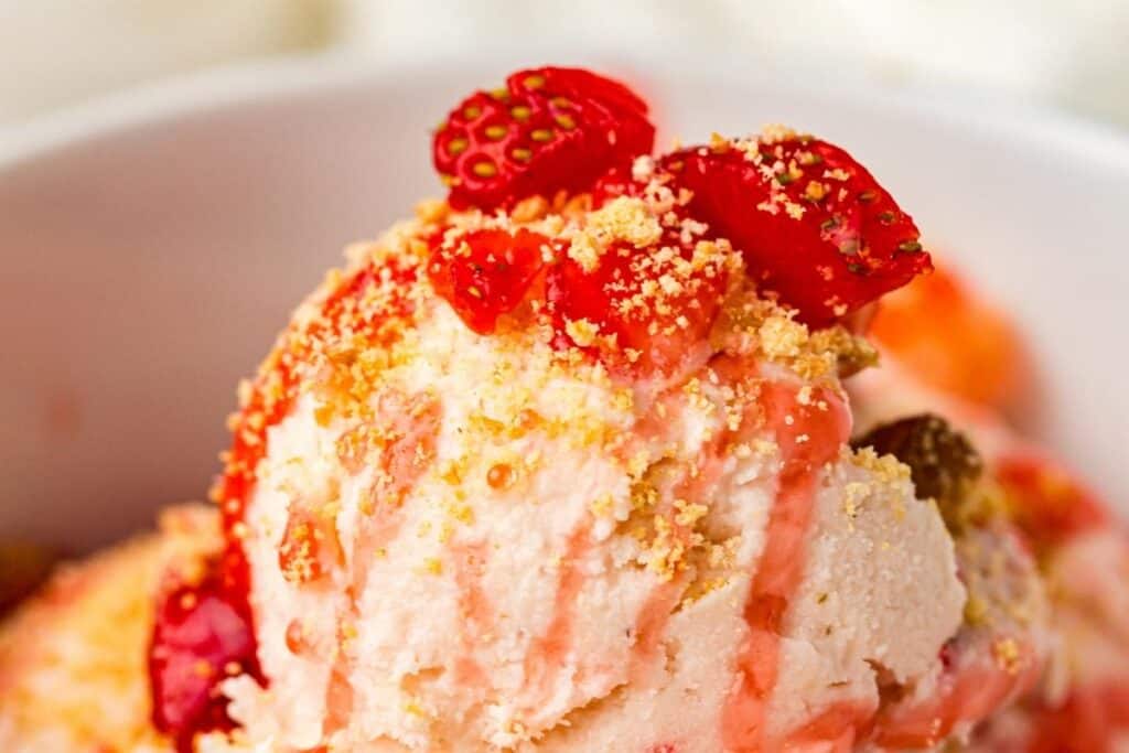 A close-up of ice cream topped with crushed crumbs, strawberry sauce, and fresh strawberry pieces in a white bowl.
