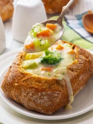 A bread bowl filled with creamy vegetable chowder, with a spoon scooping soup out; salt and pepper shakers, celery, and a wooden spoon are nearby.