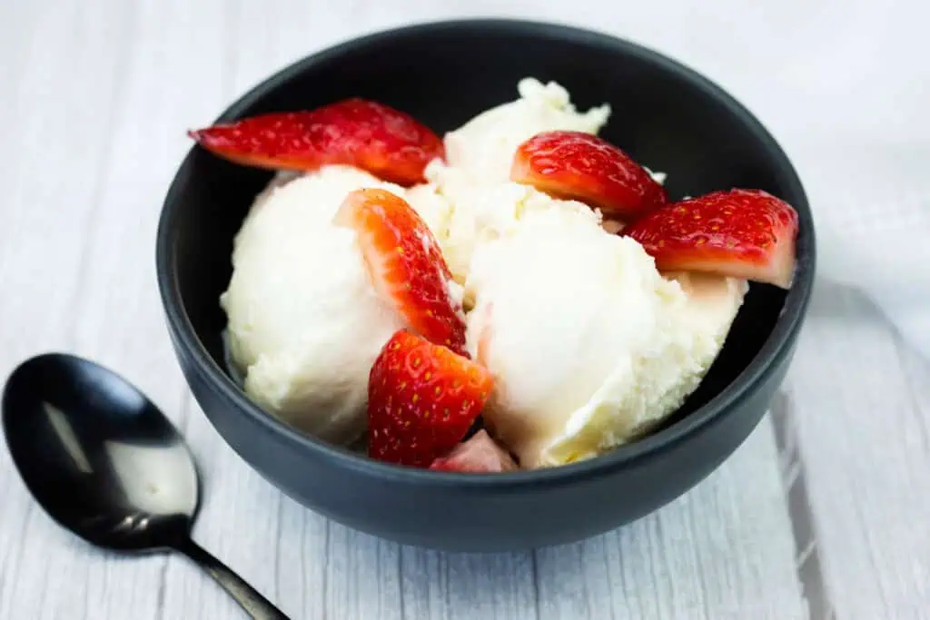 A black bowl with scoops of vanilla ice cream topped with sliced strawberries, placed on a white surface next to a black spoon.