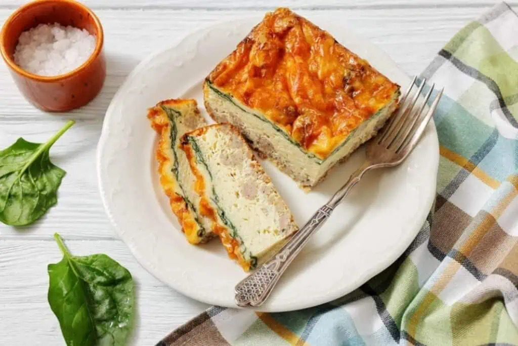 A plate with slices of baked egg casserole containing spinach and cheese, accompanied by a fork, a cup of coarse salt, and fresh spinach leaves on the side.