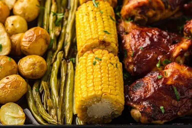 Close-up of roasted baby potatoes, green beans, corn on the cob, and barbecue chicken pieces on a tray.