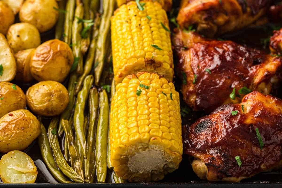 Roasted baby potatoes, green beans, corn on the cob, and barbecue chicken arranged on a sheet pan.