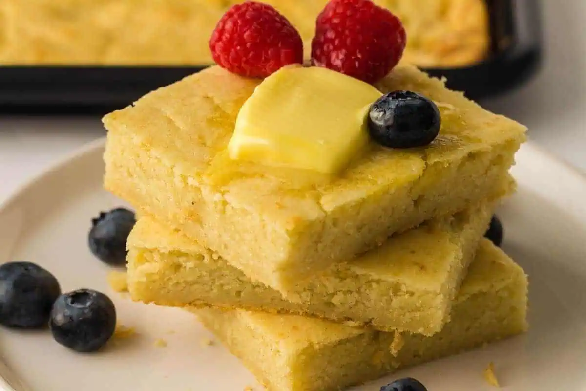 Three stacked pieces of cornbread topped with a pat of butter, raspberries, and a blueberry, with additional blueberries on the plate.