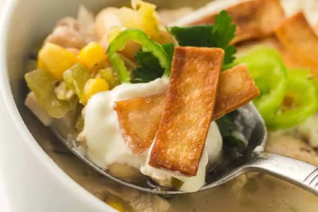 A spoonful of white chili with sour cream, corn, green chili slices, cilantro, and crispy tortilla strips.