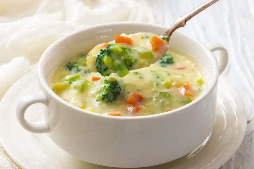 A white bowl of creamy vegetable soup with broccoli and carrots, with a spoon lifting a portion.