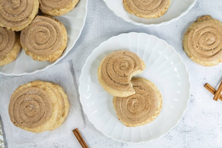 Several cinnamon swirl cookies are arranged on white plates; one cookie on the center plate has a bite taken out. Cinnamon sticks are placed nearby on a light background.