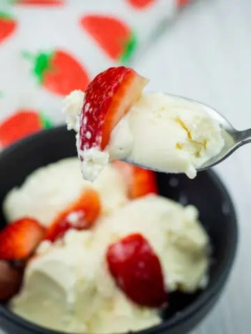A spoonful of vanilla ice cream with a strawberry slice held above a bowl containing more ice cream and strawberries.