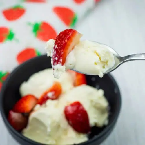 A spoonful of vanilla ice cream with a strawberry slice held above a bowl containing more ice cream and strawberries.