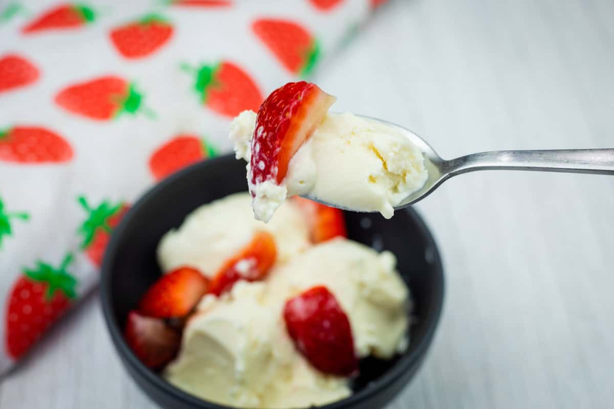 A spoonful of vanilla ice cream and a strawberry held above a bowl with more ice cream and strawberries, next to a strawberry-patterned cloth.