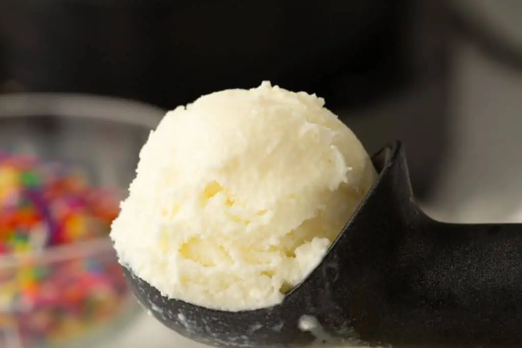 A close-up of a scoop of vanilla ice cream in a black ice cream scooper, with a blurred bowl of colorful sprinkles in the background.