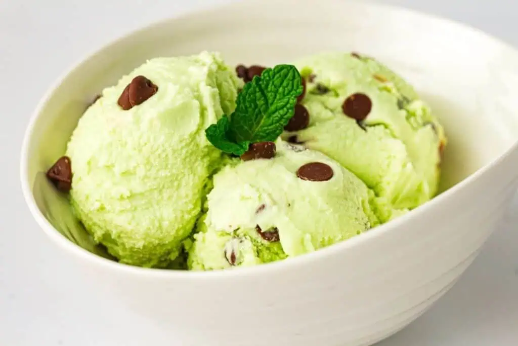 Three scoops of mint chocolate chip ice cream with chocolate chips and a mint leaf garnish in a white bowl.