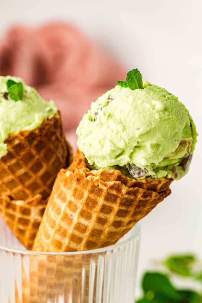 Two waffle cones filled with light green mint chocolate chip ice cream, each topped with a small mint leaf, standing in a clear glass.