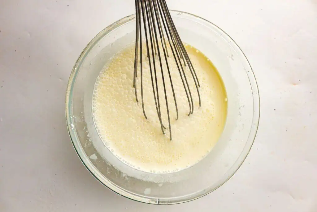 A metal whisk sits in a glass bowl filled with a pale, frothy liquid mixture on a light-colored surface.