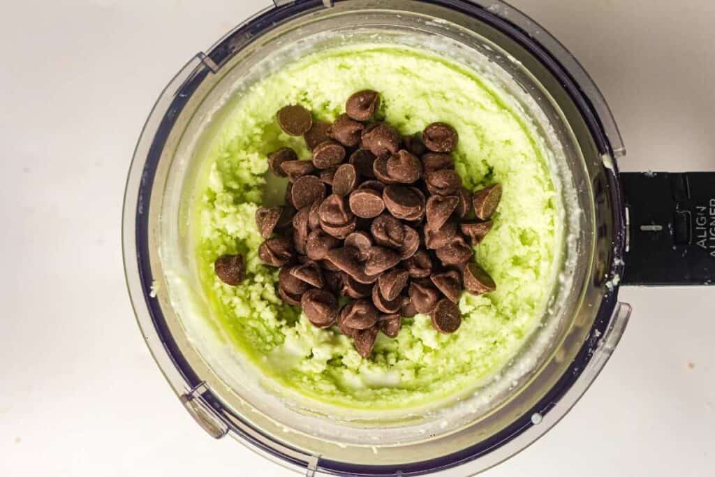 A food processor bowl containing green batter with chocolate chips added on top, viewed from above.