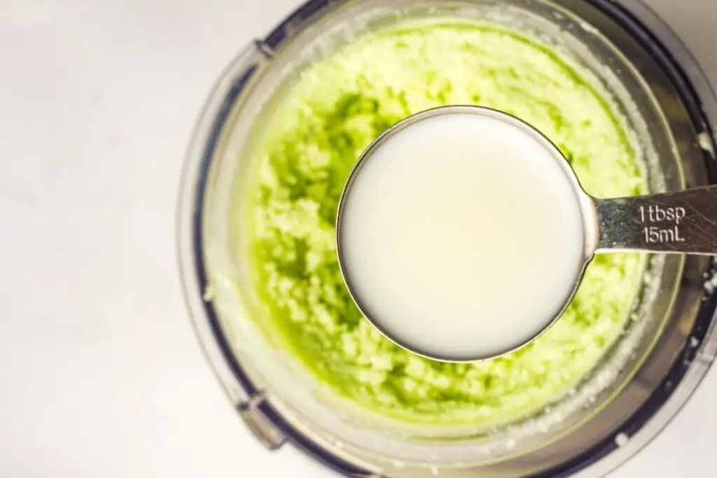 A metal tablespoon filled with milk is held above a food processor containing a green mixture.