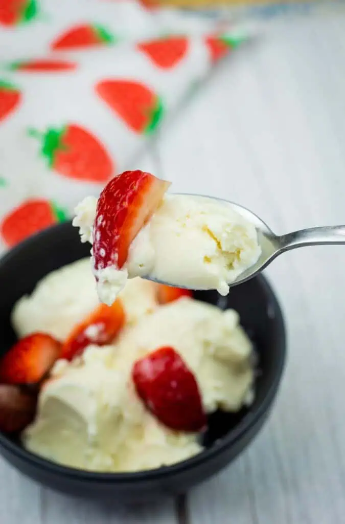 A spoonful of vanilla ice cream and a strawberry slice held above a bowl filled with ice cream and strawberries.