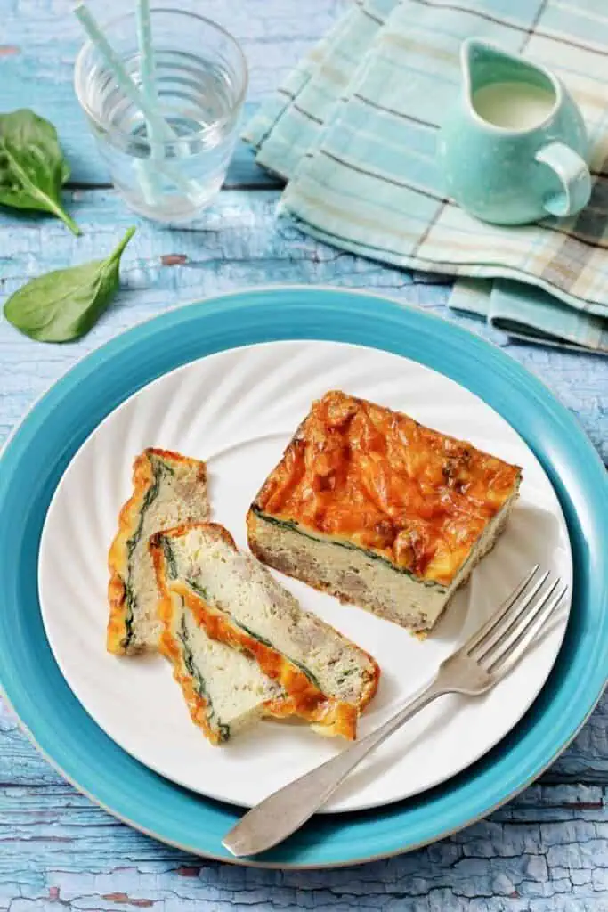 Slices of baked spinach and cheese casserole are served on a white plate with a fork, set on a blue charger, alongside a glass of water and a small pitcher.