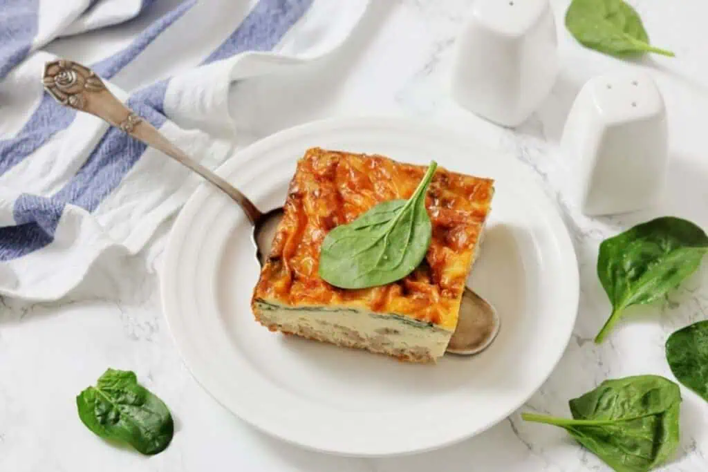 A square slice of baked casserole topped with a spinach leaf is served on a white plate with a spoon, surrounded by fresh spinach leaves and salt and pepper shakers.