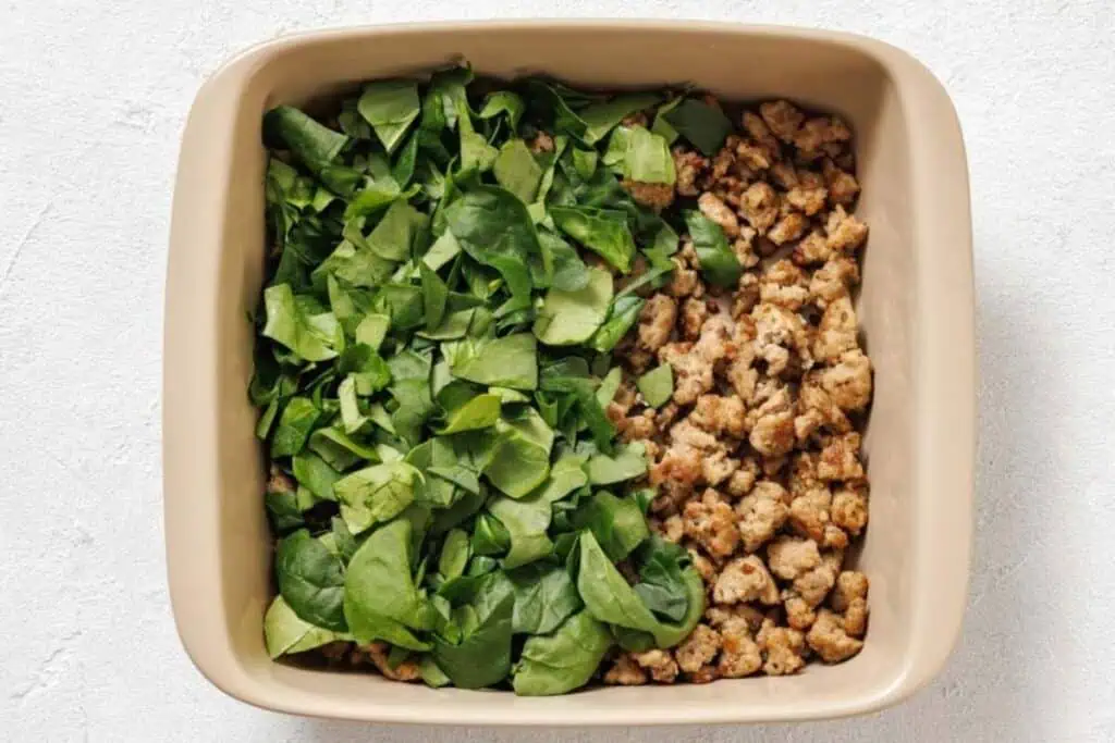 A square baking dish filled halfway with chopped spinach and halfway with cooked crumbled sausage.