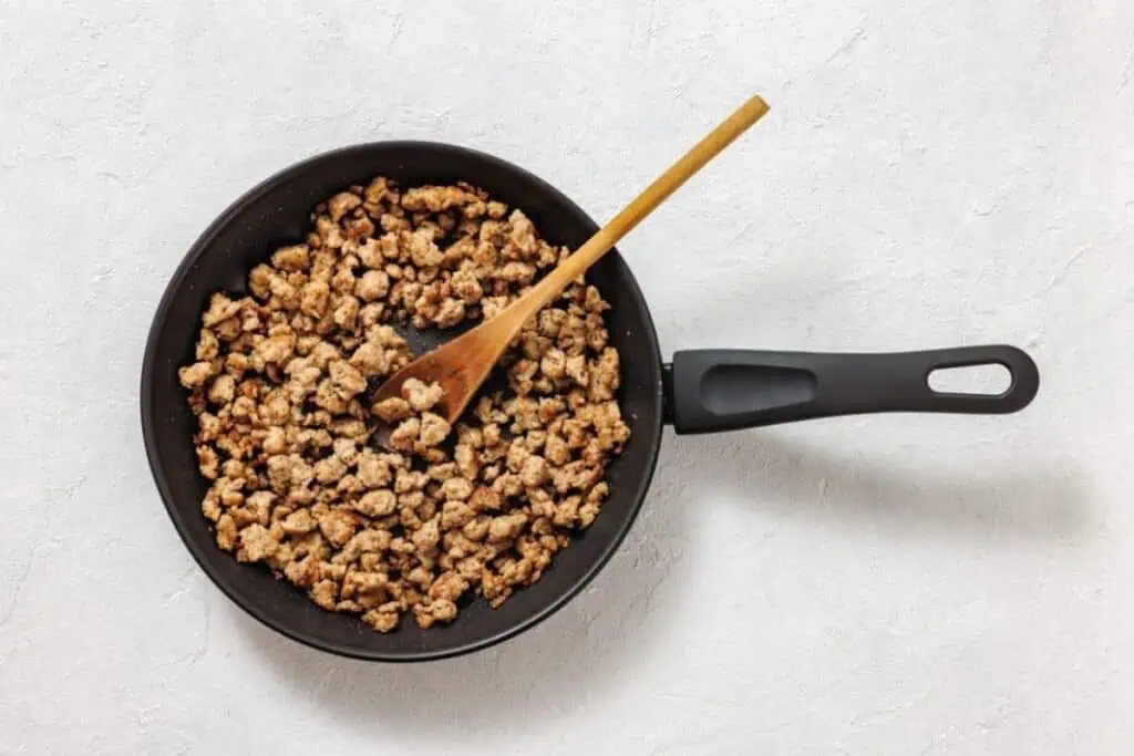 A frying pan filled with cooked ground meat and a wooden spoon on a light-colored surface.