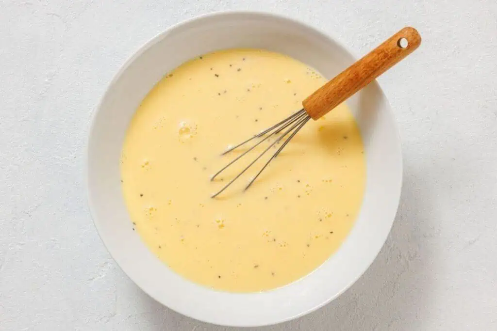 A white bowl filled with beaten eggs and black pepper, with a metal whisk featuring a wooden handle resting inside.