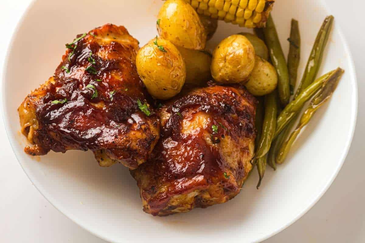 A white plate with two pieces of glazed barbecue chicken, roasted baby potatoes, corn on the cob, and green beans.