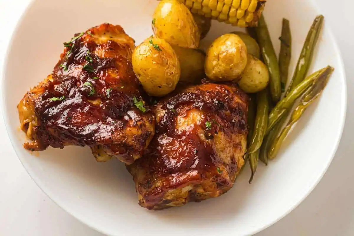A white plate with two pieces of glazed barbecue chicken, roasted baby potatoes, corn on the cob, and green beans.