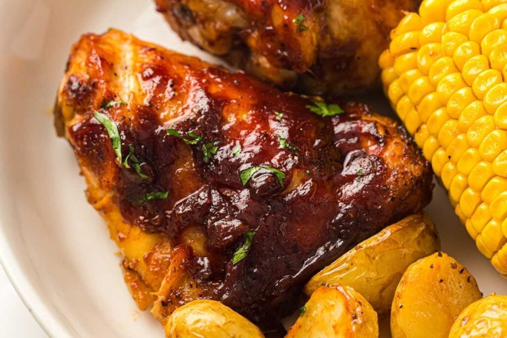 Barbecued chicken thigh with sauce, served with roasted potatoes and a piece of corn on the cob on a white plate.