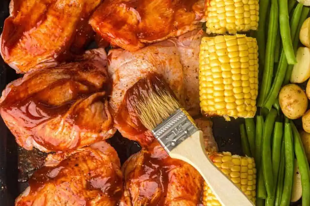 Raw chicken pieces are being brushed with barbecue sauce next to corn on the cob, green beans, and baby potatoes on a baking tray.