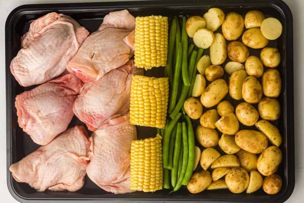 A baking sheet with raw chicken thighs, corn on the cob, green beans, and halved baby potatoes arranged in rows.